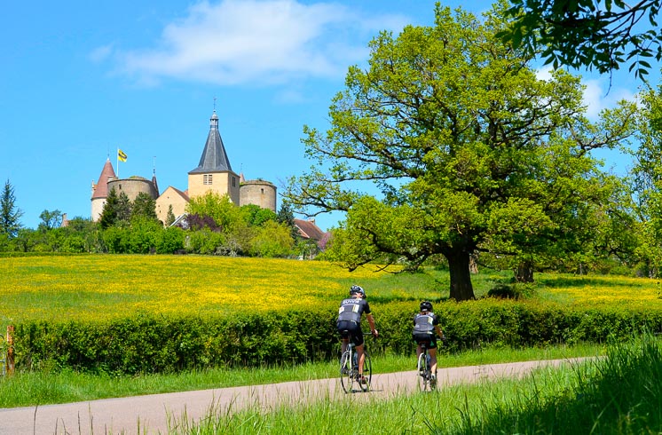 Two people riding bikes on a small road in Burgundy, surrounded by greenery and a castle in the distance