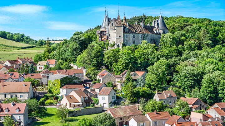 The castle at La Rochepot seen from a distance, surrounded by trees and a village of red rooftops